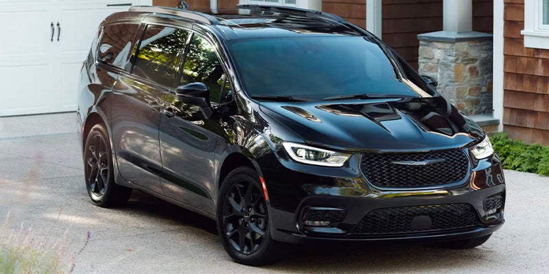 Dark gray Chrysler Pacifica parked in a residential driveway, viewed from the front with a modern house entrance in the background.