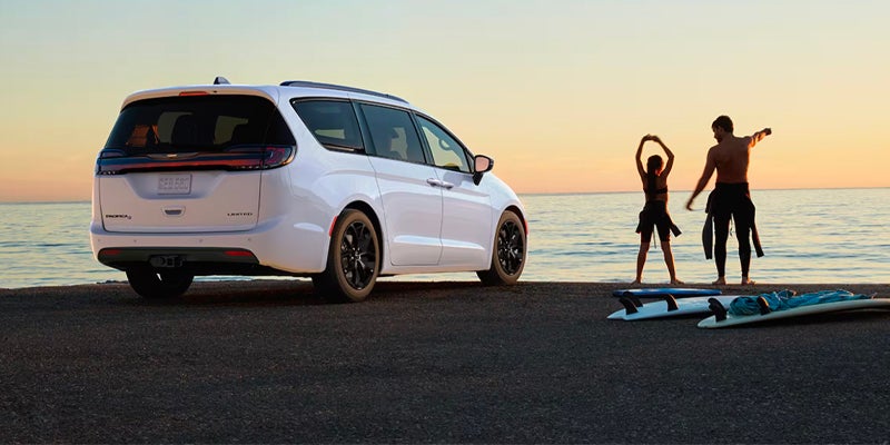 White Chrysler Pacifica minivan parked beside a waterfront at sunset, with people standing near paddleboards in the distance.