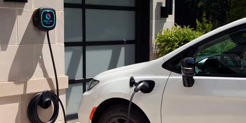 White Chrysler Pacifica Hybrid connected to a home charging station, showing the charging port and cable attached to the vehicl