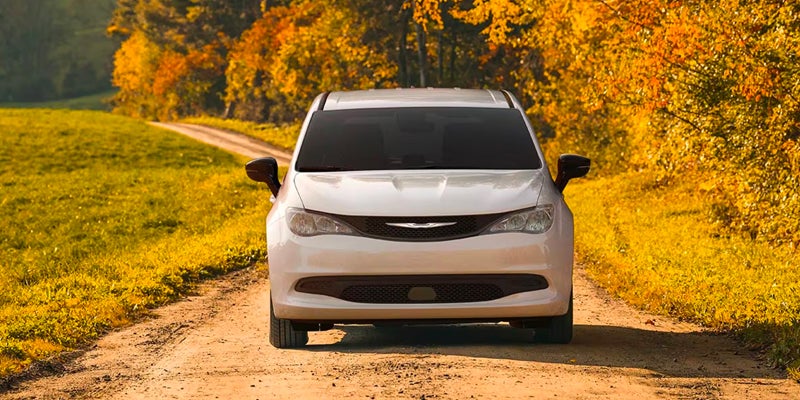 White Chrysler minivan viewed from the front, parked on a tree-lined dirt road surrounded by autumn foliage.