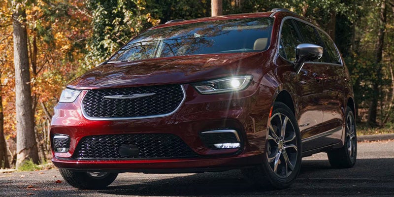 Red Chrysler Pacifica minivan parked on a paved road, shown from a front three-quarter angle with autumn trees in the background.
