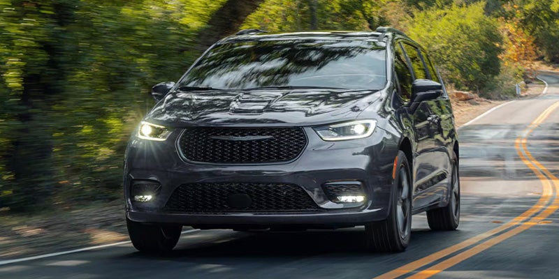 Gray Chrysler Pacifica minivan driving on a winding road through a wooded area, shown from a front three-quarter angle.