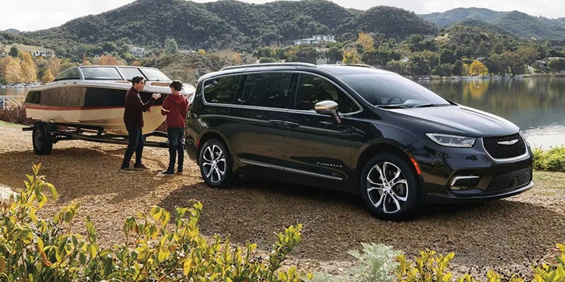 Black Chrysler Pacifica parked beside a lake with a boat trailer attached, highlighting towing capability in an outdoor recreational setting.