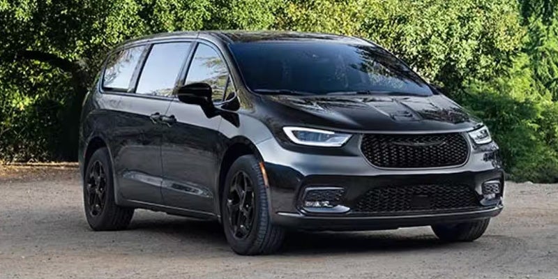 Black Chrysler Pacifica minivan parked on a gravel surface, shown from a front three-quarter angle with trees and greenery in the background