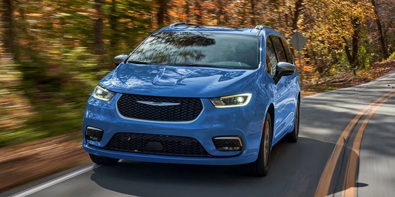 Blue Chrysler Pacifica minivan driving on a tree-lined road, shown from a front three-quarter angle in daylight.