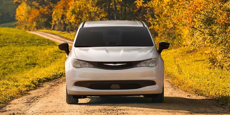 White Chrysler minivan viewed from the front on a dirt road surrounded by trees with golden autumn foliage