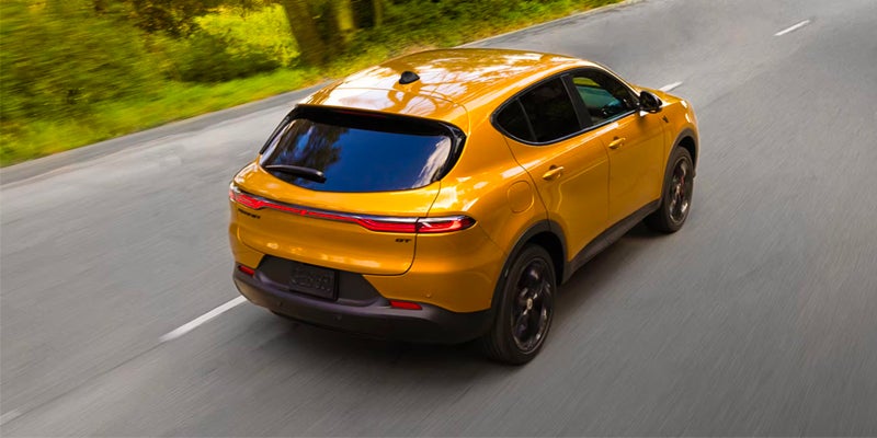 Gold Dodge Hornet compact SUV driving on a tree-lined road, shown from a rear three-quarter angle.