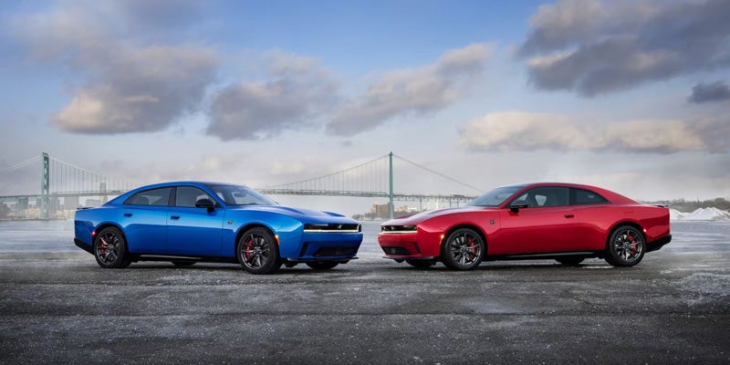 Blue and red Dodge Charger Daytona vehicles parked side by side in an open outdoor setting with a bridge visible in the background.