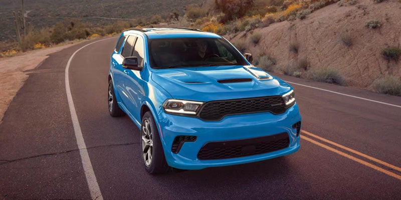 Blue Dodge Durango SUV driving on a winding road through a desert landscape, viewed from a front three-quarter angle.