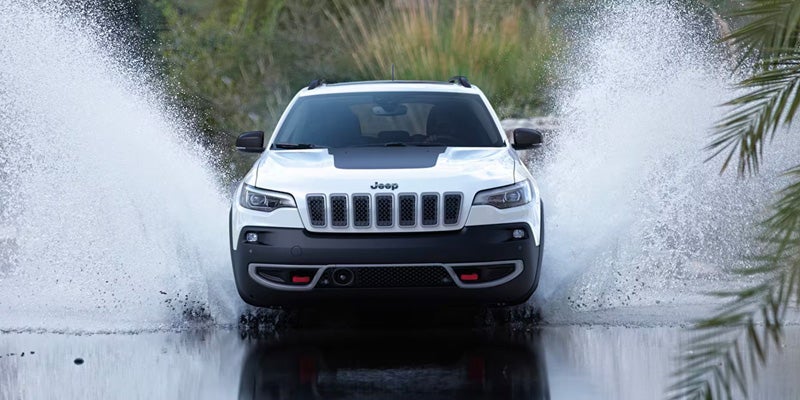 White Jeep Cherokee Trailhawk driving through shallow water, creating large splashes on both sides of the vehicle.