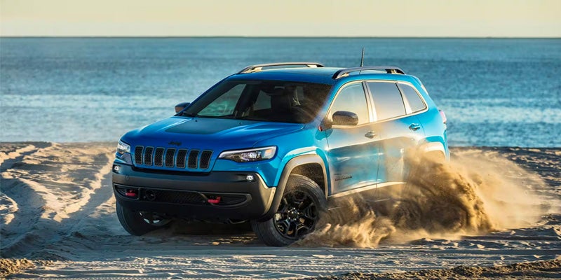 Blue Jeep Cherokee Trailhawk driving across a sandy beach near the shoreline, kicking up sand behind the vehicle.