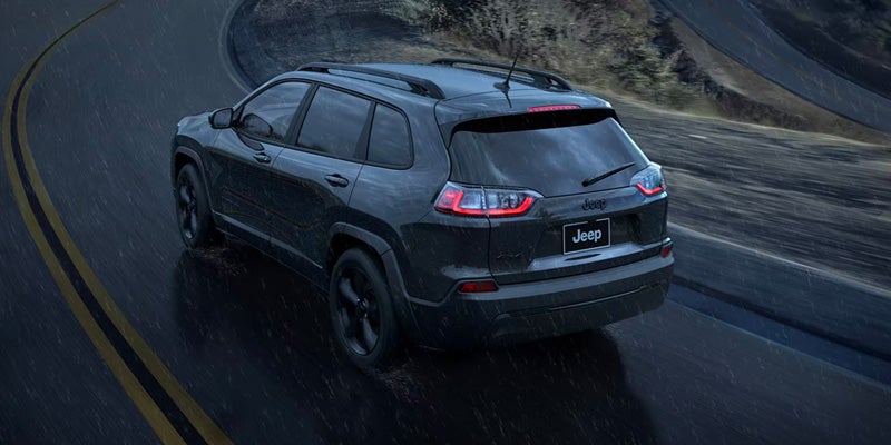 Dark-colored Jeep Cherokee driving on a wet, winding road during rainfall, viewed from the rear three-quarter angle.