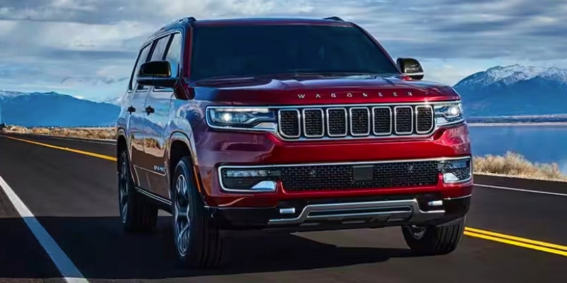 Front view of a shiny red Jeep Wagoneer SUV driving on a highway next to a lake with snowy mountains.