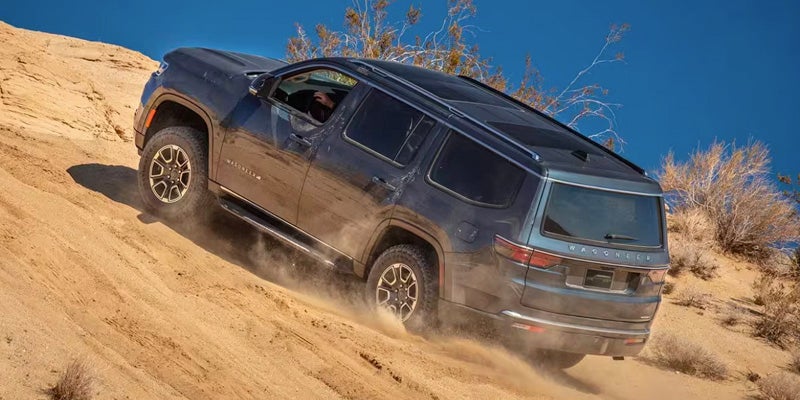 A gray Wagoneer SUV drives downhill on a sandy trail, kicking up dust.