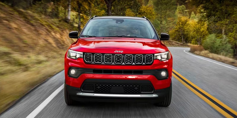 Front view of a red Jeep Compass driving on a winding road through autumn trees.