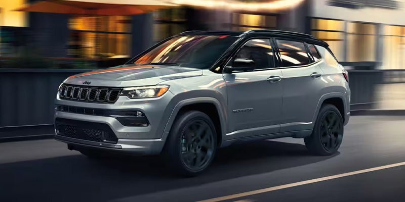 Silver Jeep Compass SUV driving on a road at dusk with city lights in the blurred background.