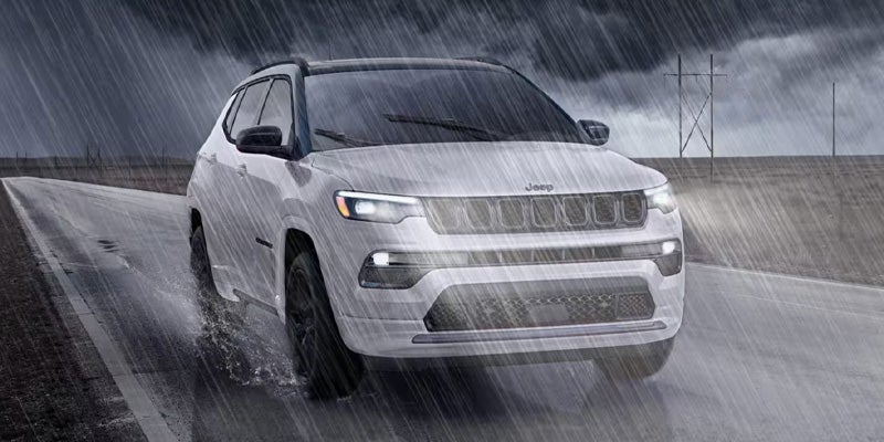 Light gray Jeep SUV driving on a wet road in heavy rain, headlights on, with utility poles in the background.