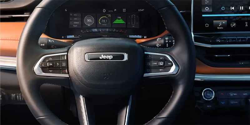 The interior of a Jeep, showing the steering wheel with controls, digital instrument cluster, and part of the infotainment screen.