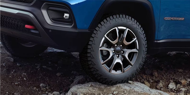 Close-up of a blue Jeep Compass on rough terrain, showcasing its front tire, alloy wheel, and part of the bumper.