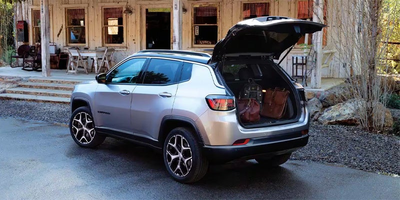 Silver Jeep Compass parked with its open trunk filled with luggage, next to a rustic wooden building.