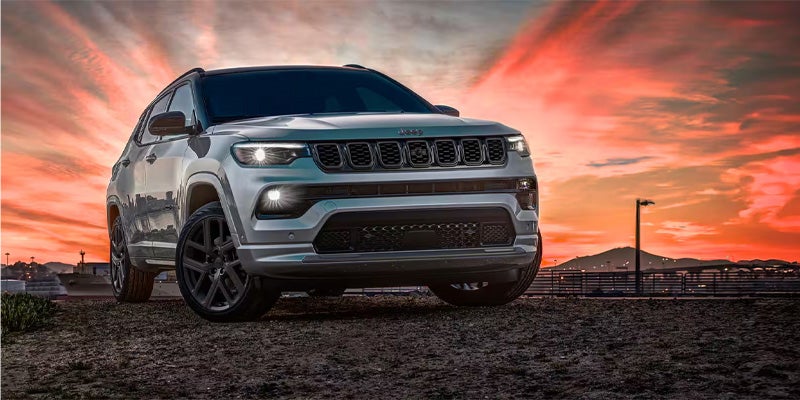 A light gray Jeep Compass SUV with headlights on, parked on dirt, with an orange and pink sunset sky.