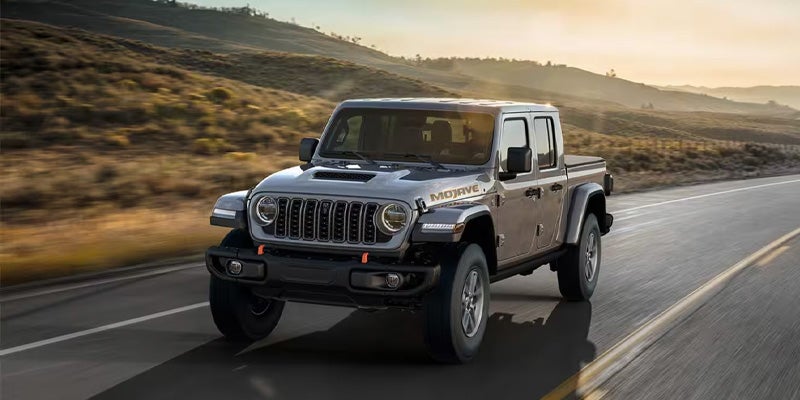 A gray Jeep Gladiator Mojave truck driving on a winding desert road at sunset with mountains in the background.