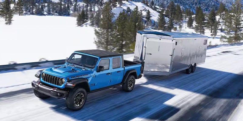 A blue Jeep Gladiator pickup truck tows a silver trailer down a snowy road, with snow-covered mountains and trees in the background.