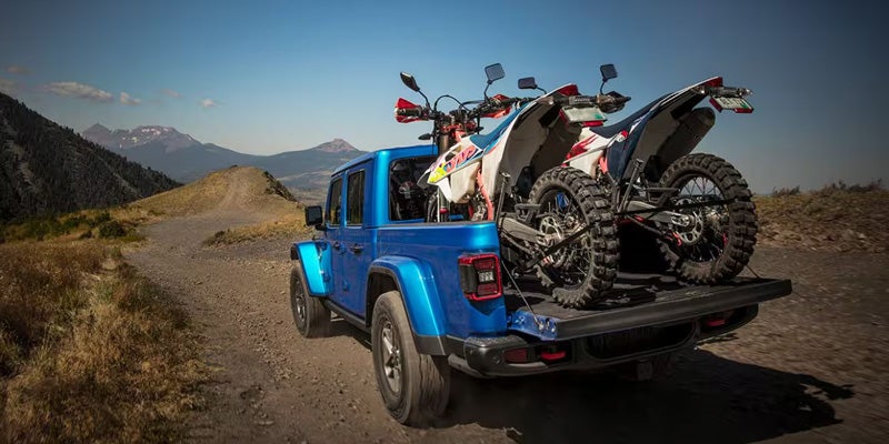 Blue Jeep Gladiator truck with two dirt bikes loaded in the bed, driving on a dirt road through mountains.