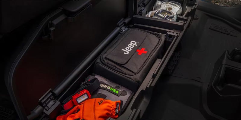 A black Jeep-branded first-aid kit, orange gloves, and other recovery gear stored in a vehicle's cargo compartment.
