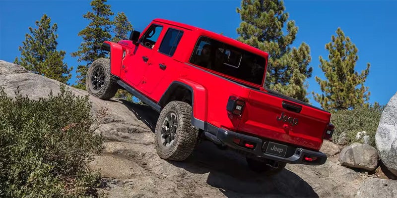 A red Jeep Gladiator pickup truck, viewed from the rear passenger side, climbing a rocky, dirt hill.
