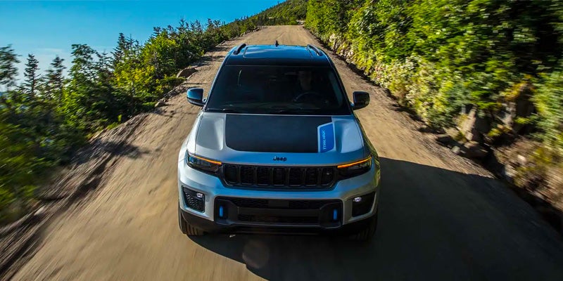 Silver Jeep Grand Cherokee 4xe with a black hood driving on a dirt road through a forest.