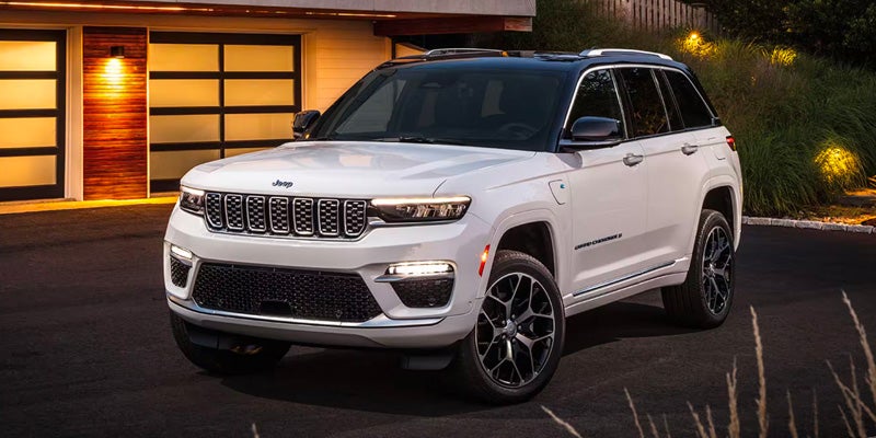 A white Jeep Grand Cherokee L 4xe parked in a driveway at dusk with the garage and landscaping lit by warm lights.