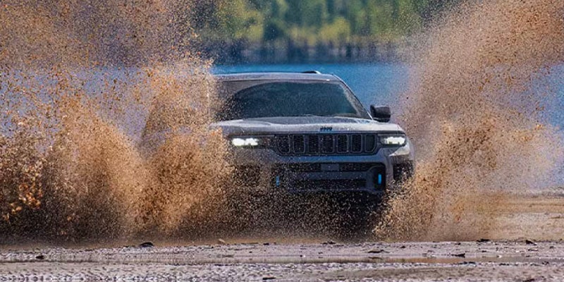 A dark SUV drives through a shallow body of water, spraying mud and water into the air.
