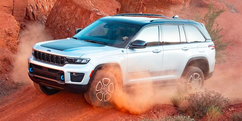 A white and black Jeep Grand Cherokee 4xe drives on a dusty red dirt road between red rock formations.