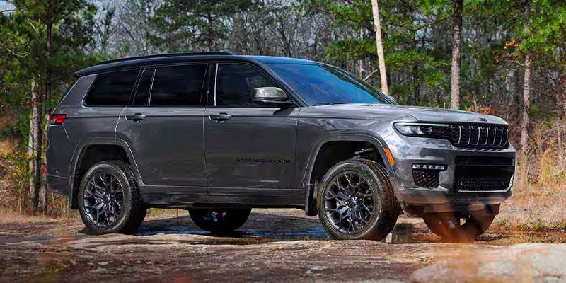A dark gray Jeep Grand Cherokee L in a wooded area with bare trees, parked on a dirt path with small rocks.