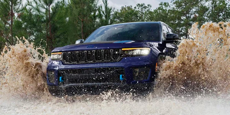 Dark blue Jeep SUV with a black grille driving through muddy water that splashes high on both sides.