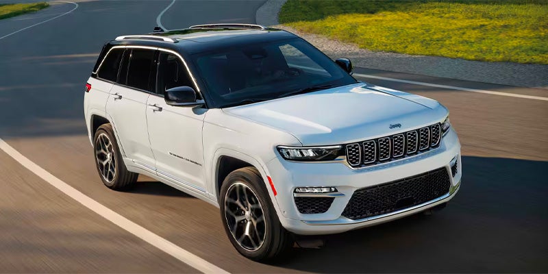 White Jeep Grand Cherokee with a black roof drives on a winding road with grass on the right.