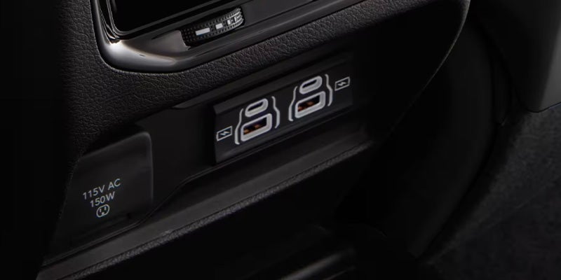 A car interior with a 115V AC 150W outlet and two USB charging ports, one USB-A and one USB-C.