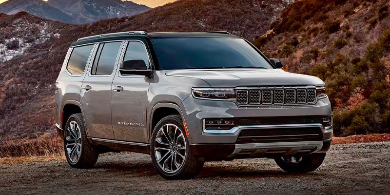 A gray Grand Wagoneer SUV with a black roof and shiny wheels is parked on a dirt road in front of mountains.