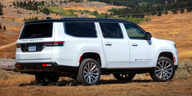 Rear 3/4 view of a white Grand Wagoneer SUV parked on a brown grassy hill, with more hills in the background.