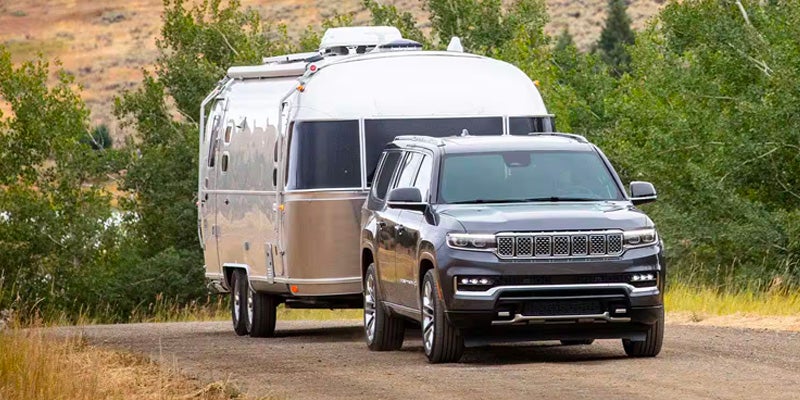 Dark gray SUV pulling a silver Airstream camper down a dirt road with green trees and hills in the background.