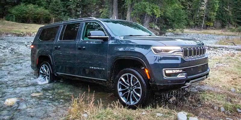 A dark gray Wagoneer SUV is half-submerged in a shallow river, with trees and brush in the background.