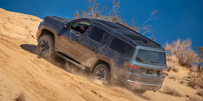 Dark gray Jeep Wagoneer driving up a steep, sandy dune, with dust kicking up from the tires, under a clear blue sky.