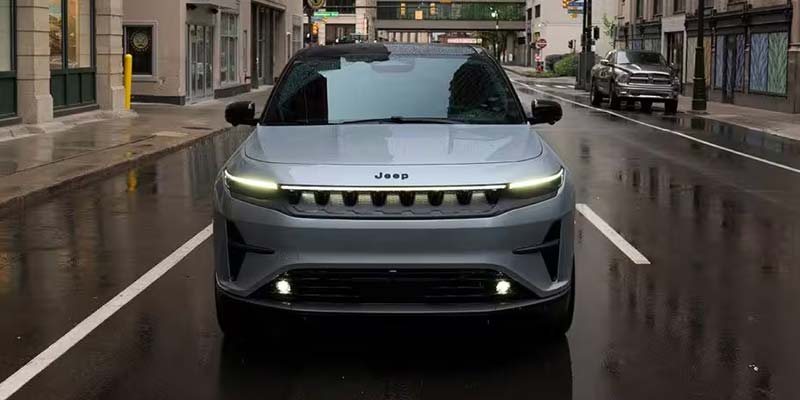 Front view of a silver Jeep parked on a wet city street, with buildings visible in the background.
