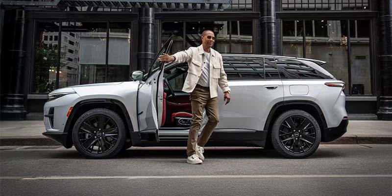 Man in a cream jacket and brown pants steps out of a white SUV with the door open, gesturing with his hand.