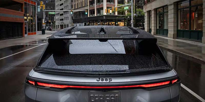 Rear view of a silver Jeep parked on a wet city street, buildings and streetlights visible in the background.