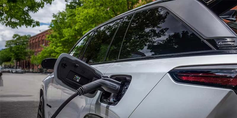 A white electric vehicle charging on a city street, with the charging cable plugged into the car's side.