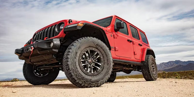A low-angle shot of a red Jeep Wrangler Rubicon on a dirt path, with mountains and cloudy sky in the background.