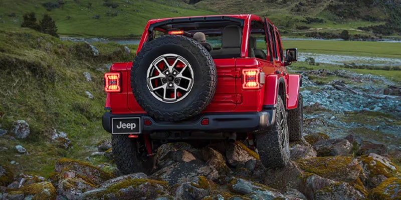 Rear view of a red Jeep Wrangler driving uphill over moss-covered rocks in a green, mountainous landscape.
