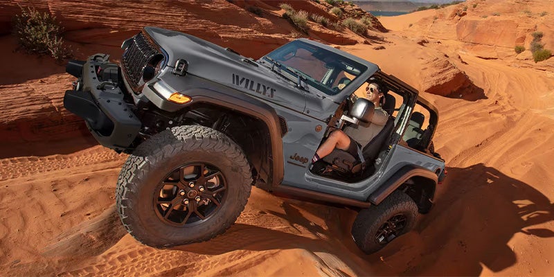 A gray Jeep Wrangler "Willys" with a passenger is off-roading up a steep red sand dune.